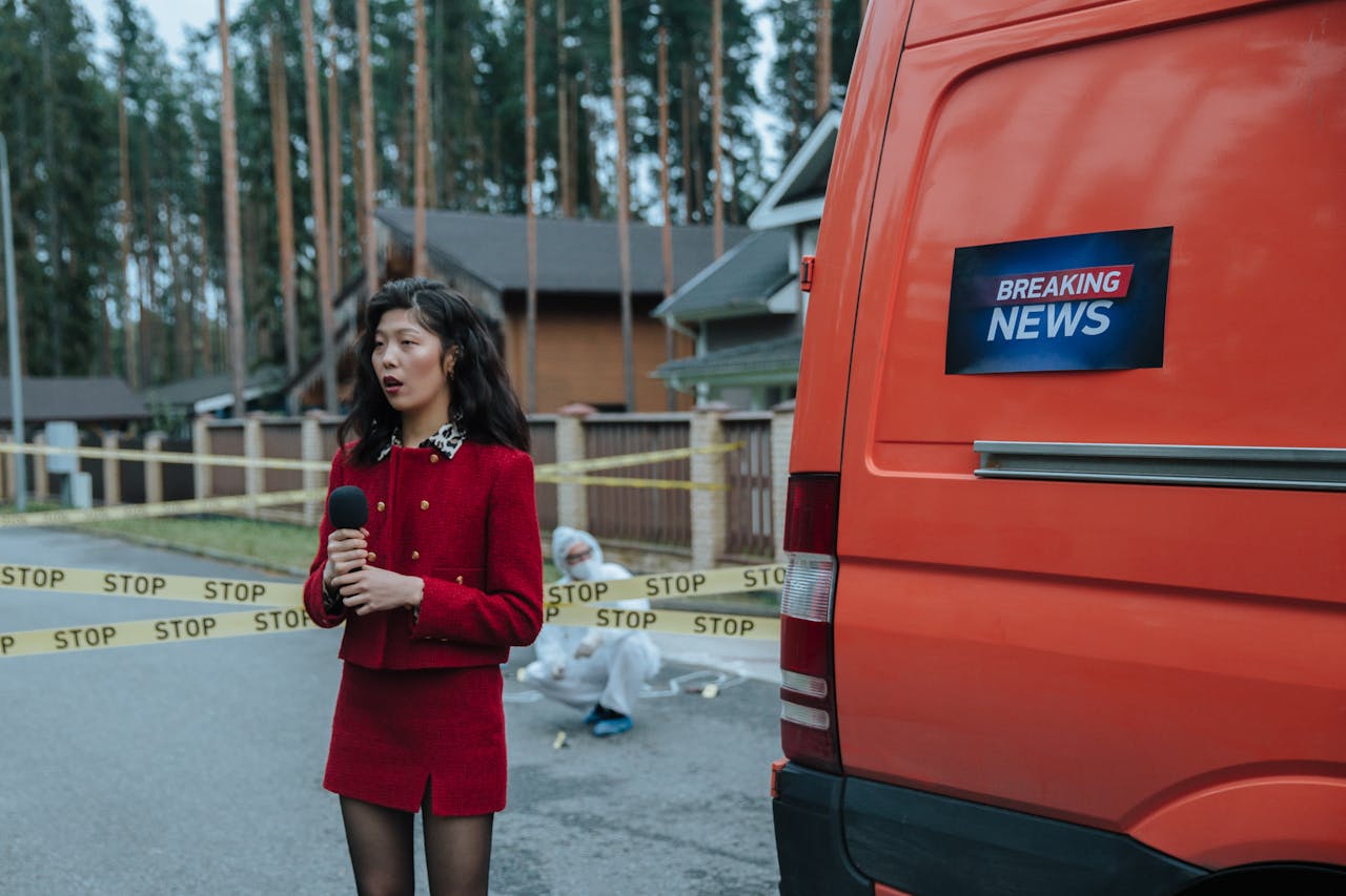 contact-img Female reporter in red outfit at an outdoor crime scene with warning tape and a news van.
